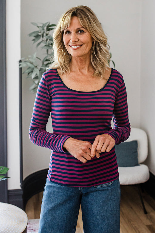 A woman standing and smiling with her hands clasped in front of her, wearing Yala Avril Scoop Neck Long Sleeve Bamboo Top in Berry Classic Stripe