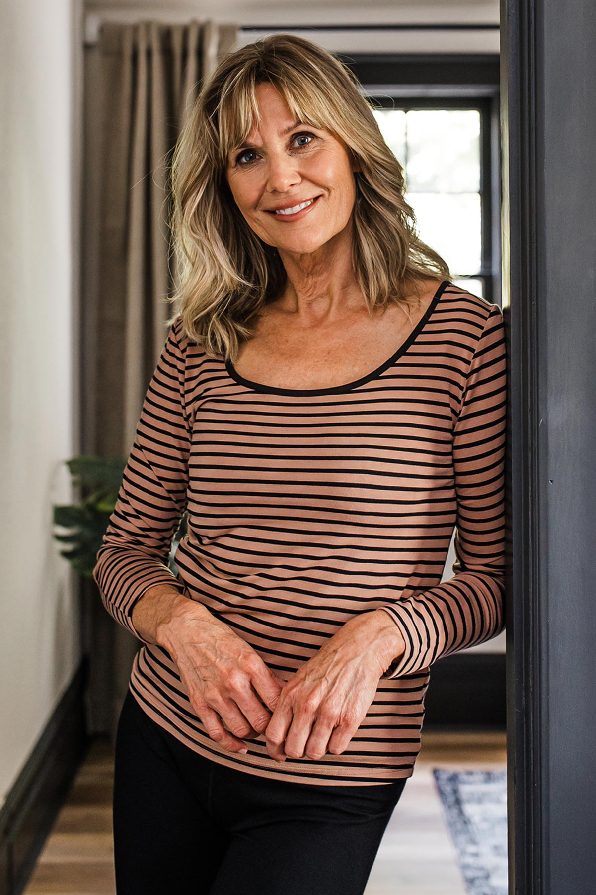 A woman standing and leaning aganst a door framewith her hands held in front of her, wearing Yala Avril Scoop Neck Long Sleeve Bamboo Top in Black Newport Stripe