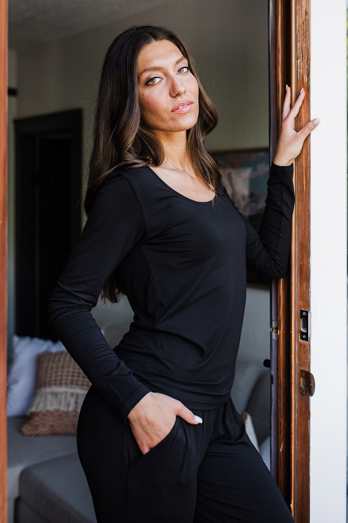 A woman standing facing to the side, one hand in her pocket adnd the other hand against a doorframe, wearing Yala Avril Scoop Neck Long Sleeve Bamboo Top in Black