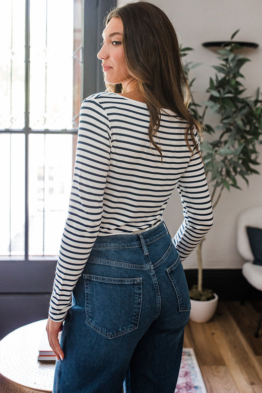 A woman standing with her back to the camera, her head turned to look back over her shoulder, wearing Yala Avril Scoop Neck Long Sleeve Bamboo Top in Navy Newport Stripe