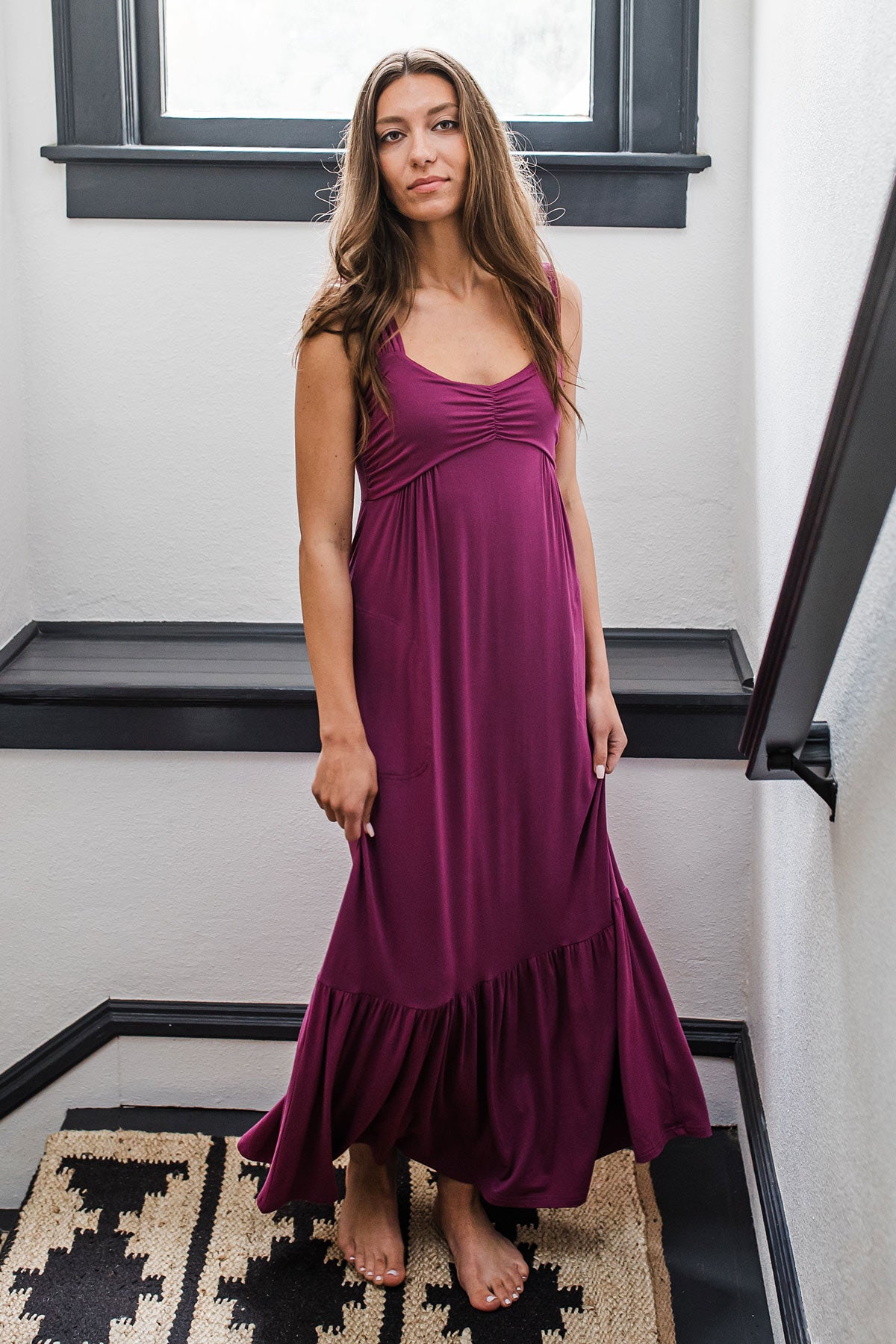 A woman standing in the landing of a stairwell and looking off to the side, wearing Yala Cleo Bamboo Babydoll Maxi Dress in Boysenberry
