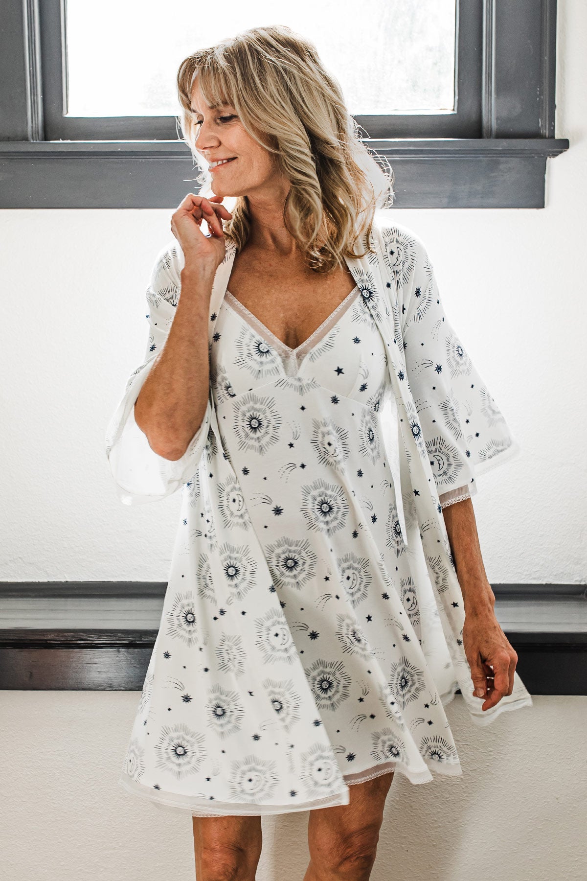 A woman standing in front of a window and smiling while looking off to the side, wearing Yala Iris Kimono Sleeve Belted Bamboo Lace Robe in Celestial Print