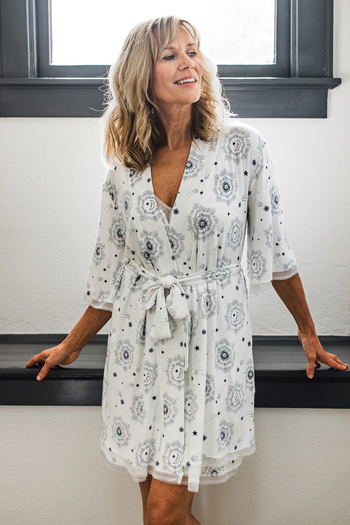 A woman leaning back against a ledge in front of a window and looking off to the side, wearing Yala Iris Kimono Sleeve Belted Bamboo Lace Robe in Celestial Print