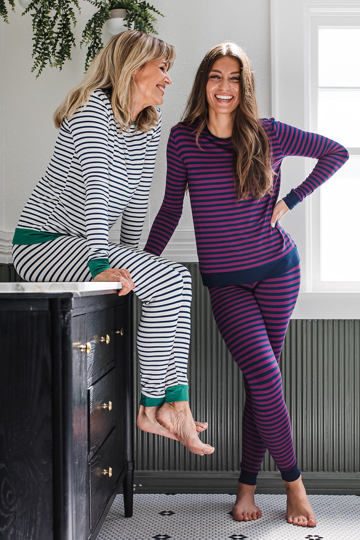 Two women smiling together, one standing with one hand on her waist and the other resting on a countertop, the other sitting upon the countertop, both wearing Yala Lola Striped Bamboo Pajama Sets, one in Berry Classic Stripe and the other in Navy Newport Stripe