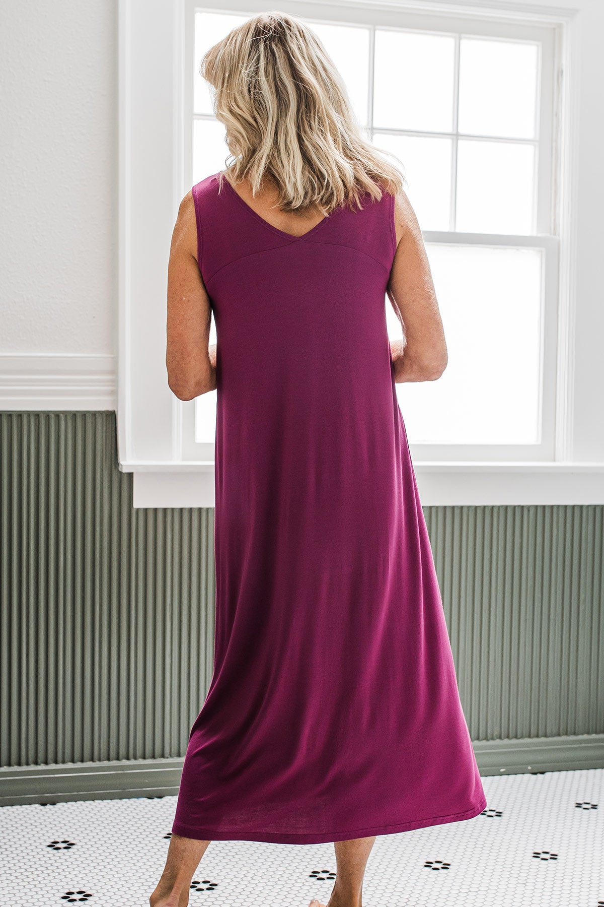 A woman standing in front of a window while facing away from the camera, wearing Yala Molly Sleeveless Bamboo Nightgown in Boysenberry