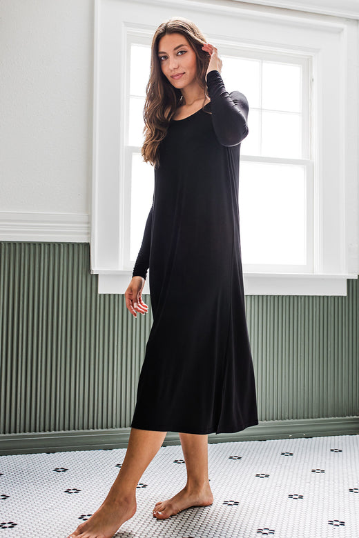A woman standing facing to the side with one foot extended, wearing Yala Scoop Neck Long Sleeve Bamboo Nightgown in Black