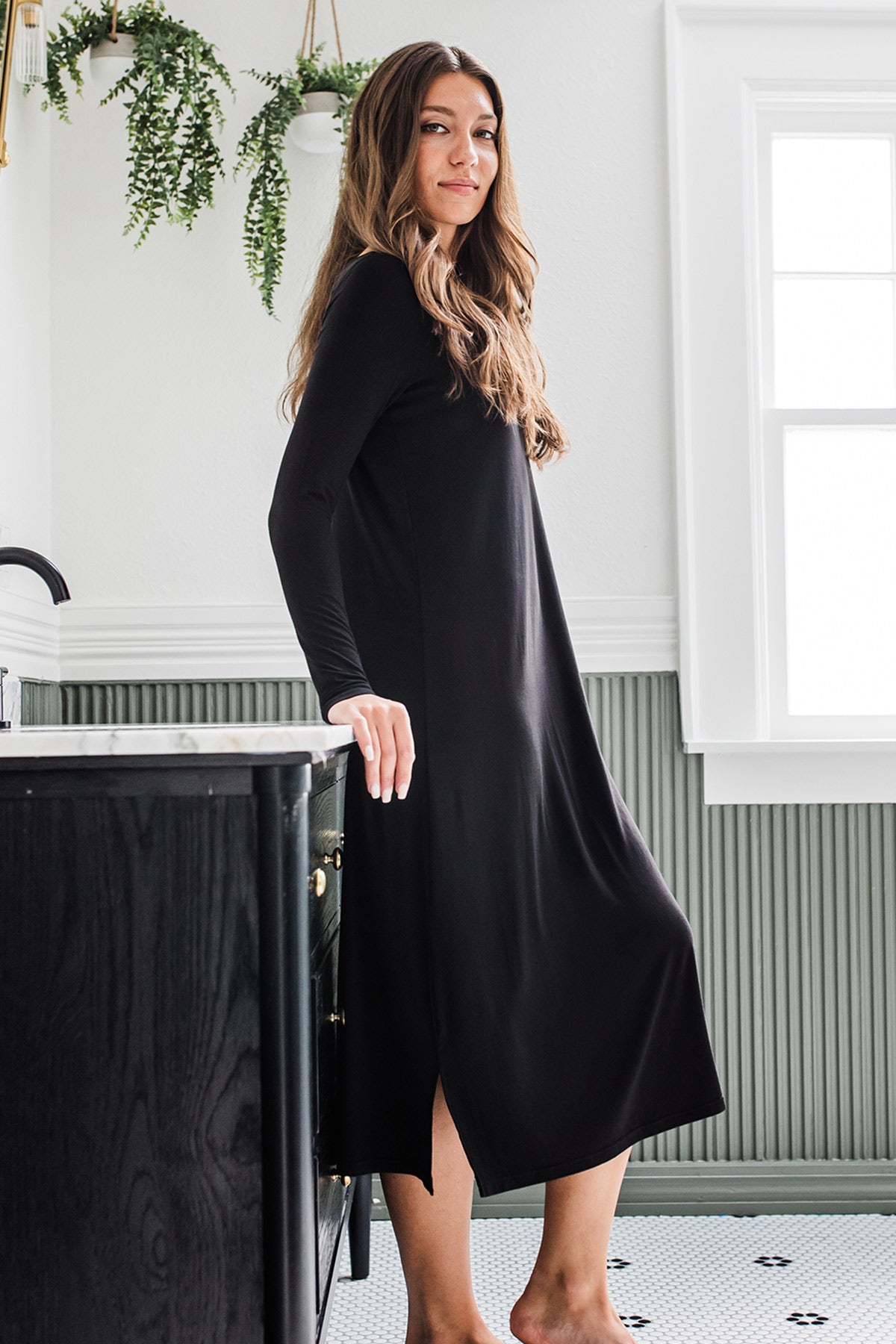 A woman standing and leaning back aganst a countertop, on knee lightly raised, wearing Yala Scoop Neck Long Sleeve Bamboo Nightgown in Black