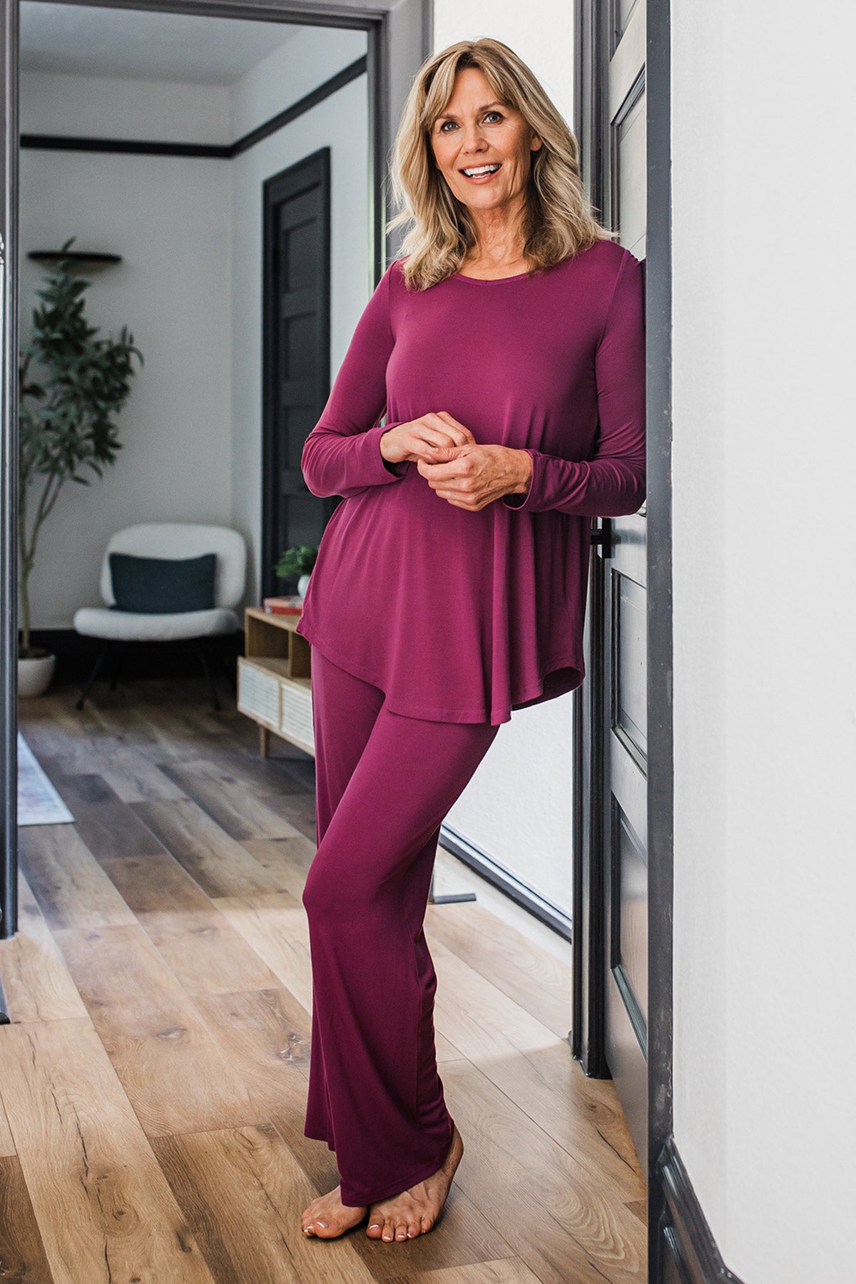 A woman standing leaning against a dorframe with her hands clasped in front of her, wearing Yala Norah Long Sleeve Bamboo Pajama Set in Boysenberry