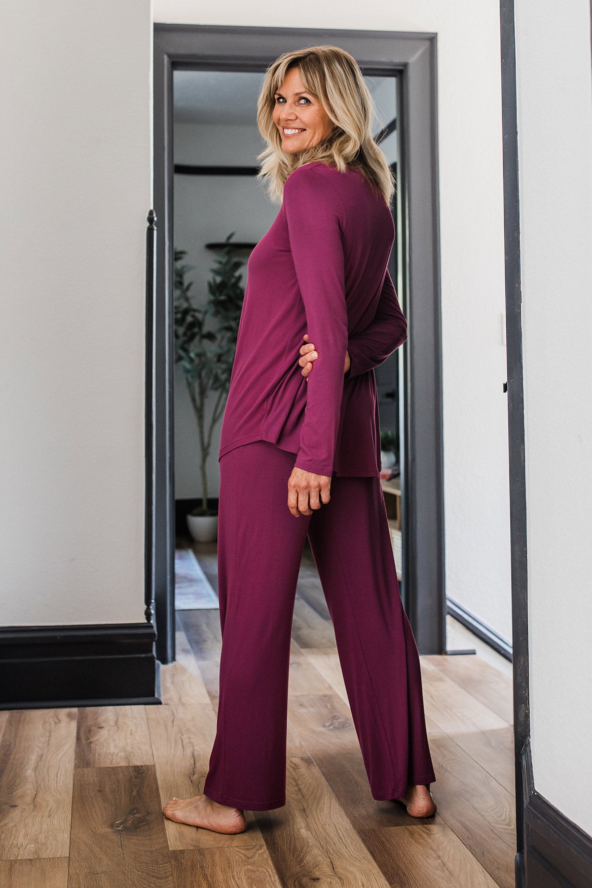 A woman standing facing away from the camera with her arms behind her back while looking back and smiling, wearing Yala Norah Long Sleeve Bamboo Pajama Set in Boysenberry