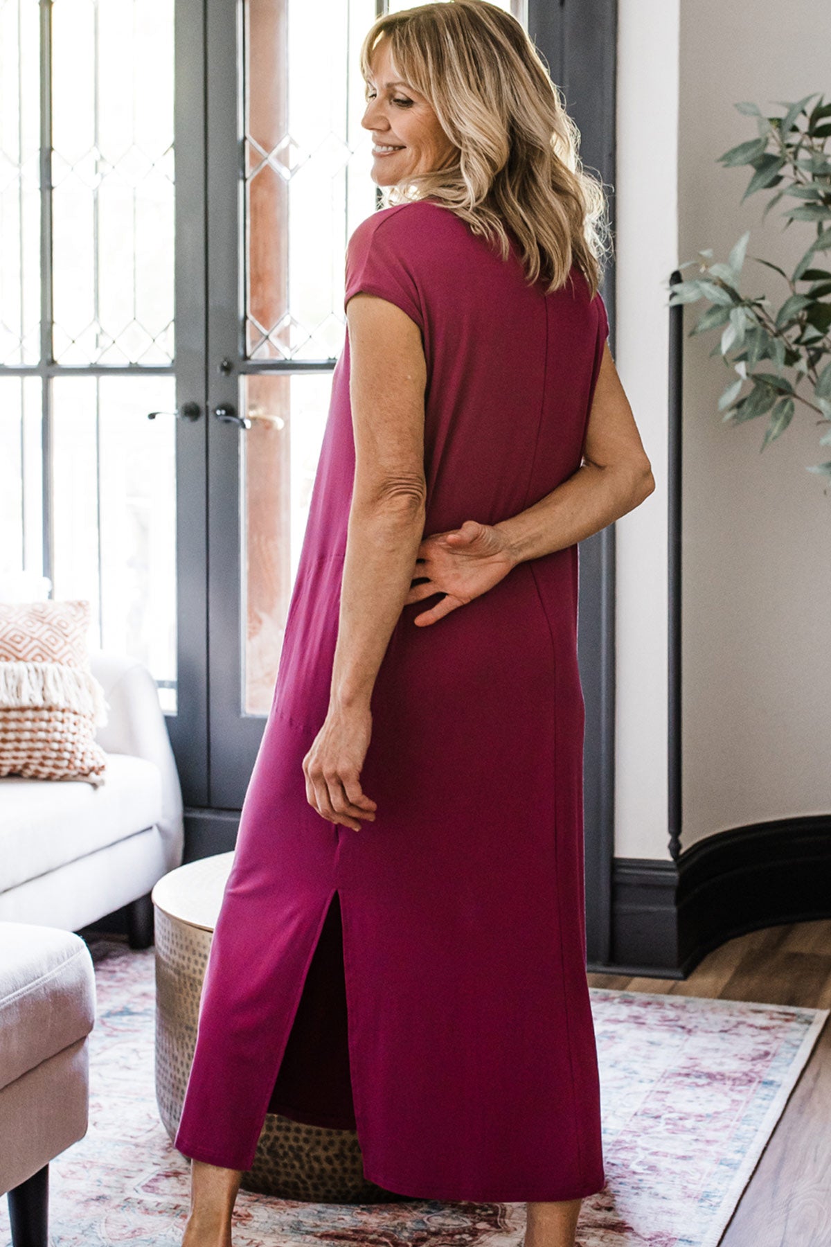 A woman standing facing away from the camera with her arms behind her back while lsmiling back over her shoulder, wearing Yala Sloane V-Neck Cap Sleeve Bamboo Maxi Dress in Boysenberry