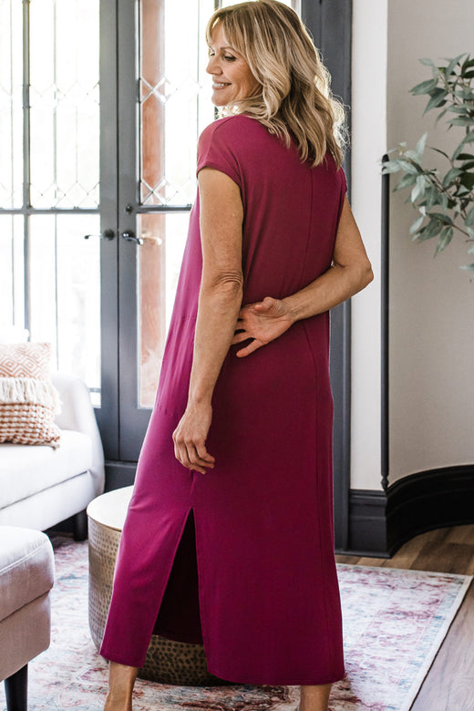 A woman standing facing away from the camera with her arms behind her back while lsmiling back over her shoulder, wearing Yala Sloane V-Neck Cap Sleeve Bamboo Maxi Dress in Boysenberry