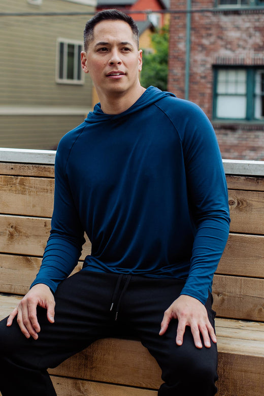 A man sitting on a bench outside with his hands on his knees, looking off to the side, wearing Yala Men's Travis Hooded Long Sleeve Raglan Bamboo Tee Shirt in Navy