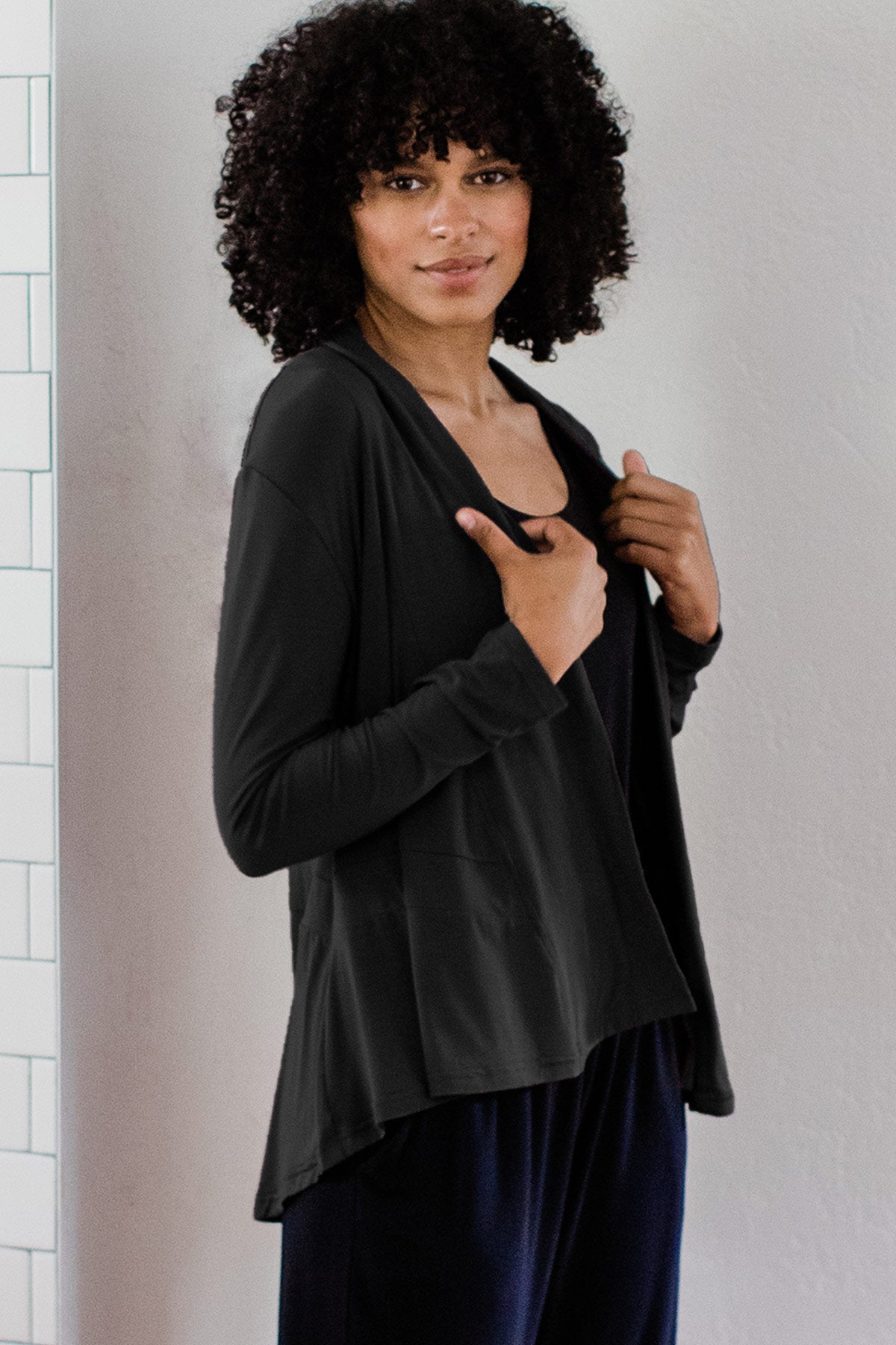 A woman standing facing to the side, both her hands holding the hem of her wrap, wearing Yala Tatum Hi-Low Bamboo Cardigan Wrap in Black