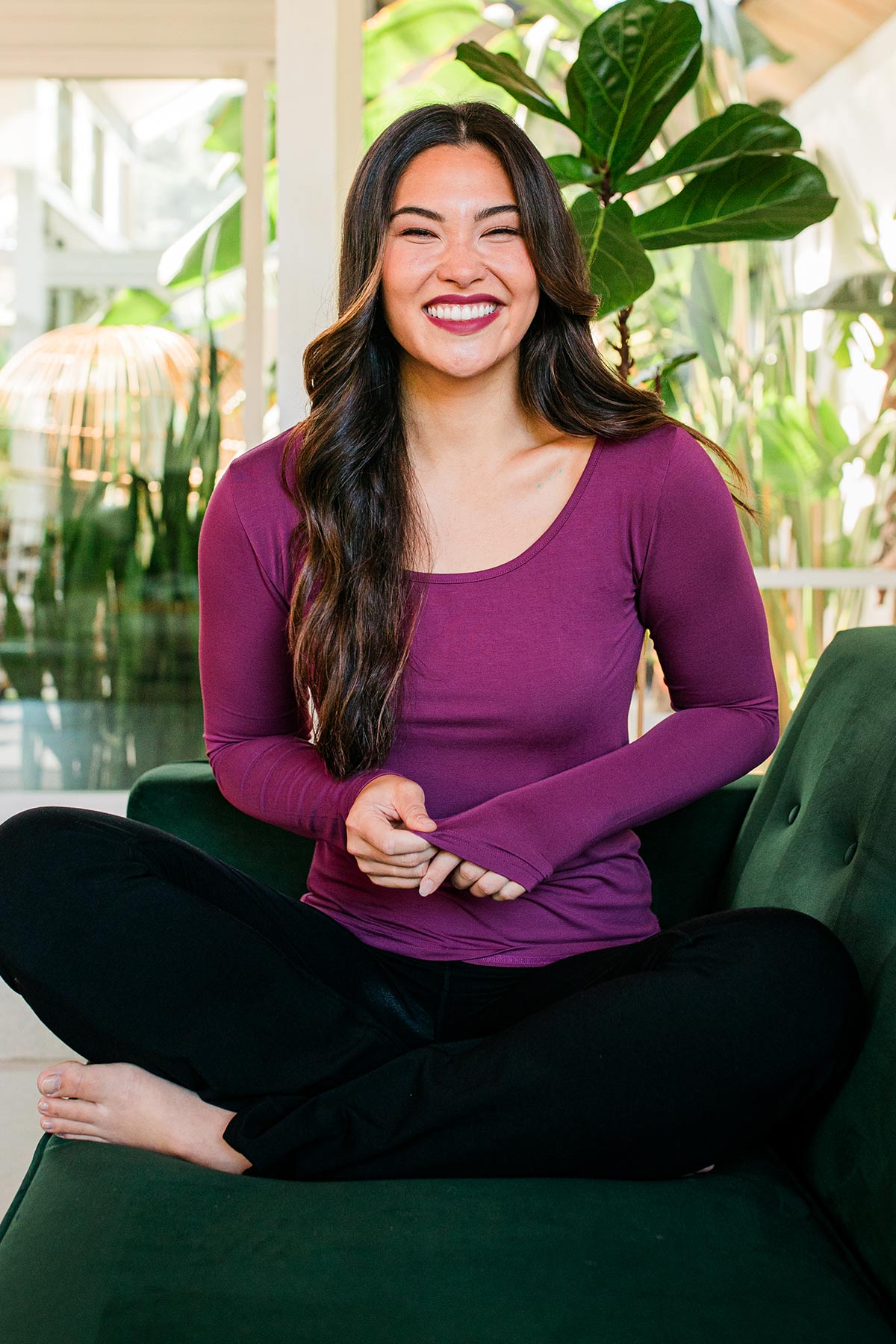 A woman sitting on a couch with her legs crossed, a smile on her face, wearing Yala Avril Scoop Neck Long Sleeve Bamboo Top in Boysenberry