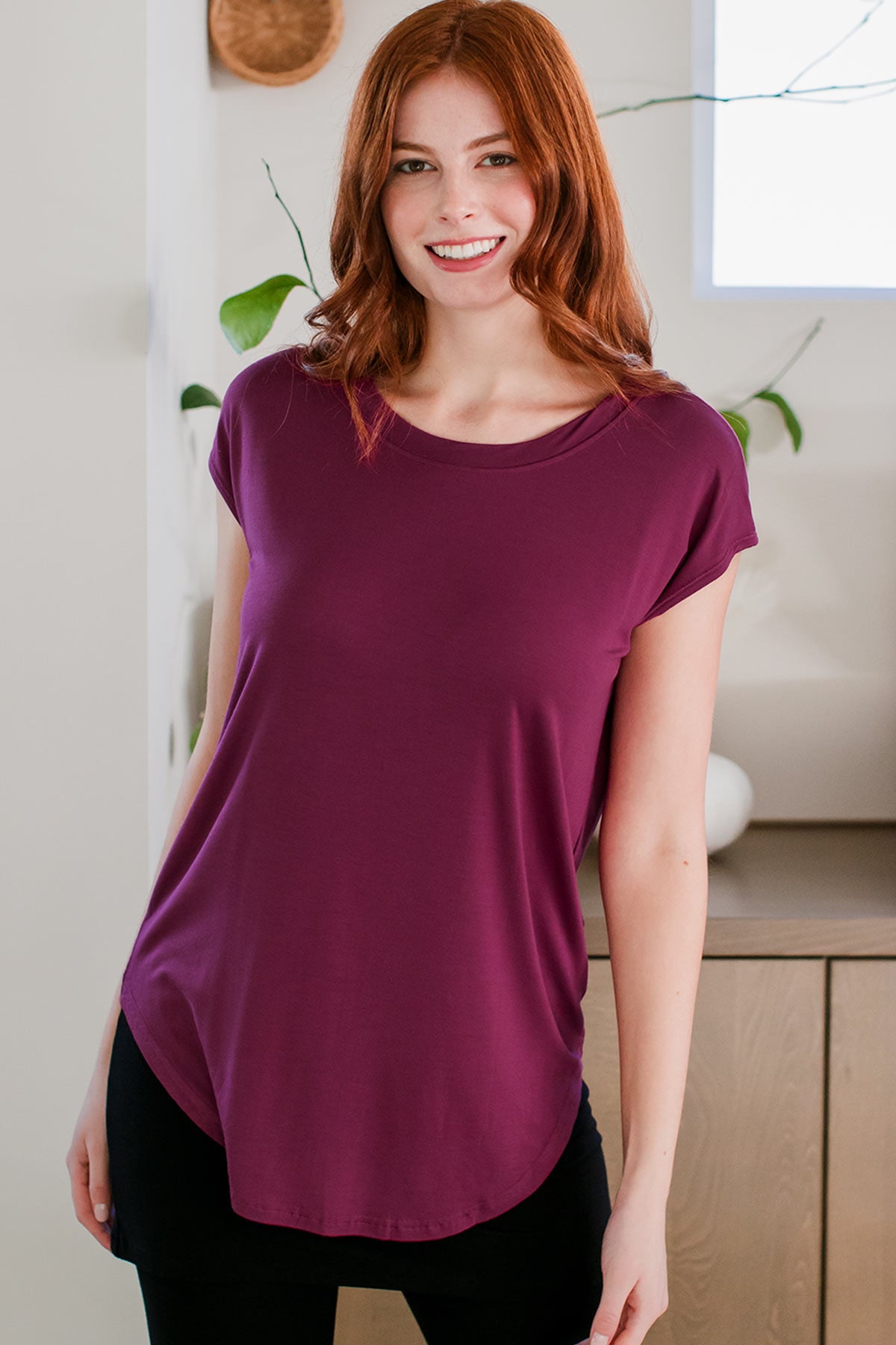 A woman standing with her hips tilted to the side and a smile on her face, wearing Cassidy Cap Sleeve Bamboo Boyfriend Tunic in Boysenberry