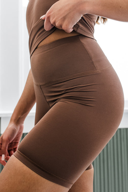 Close up shot of a woman's hips and waist, the hem of her shirt slightly lifted to showcase the waistband, wearing Yala Gwen ButterSoft Slip Shorts in Deep Nude