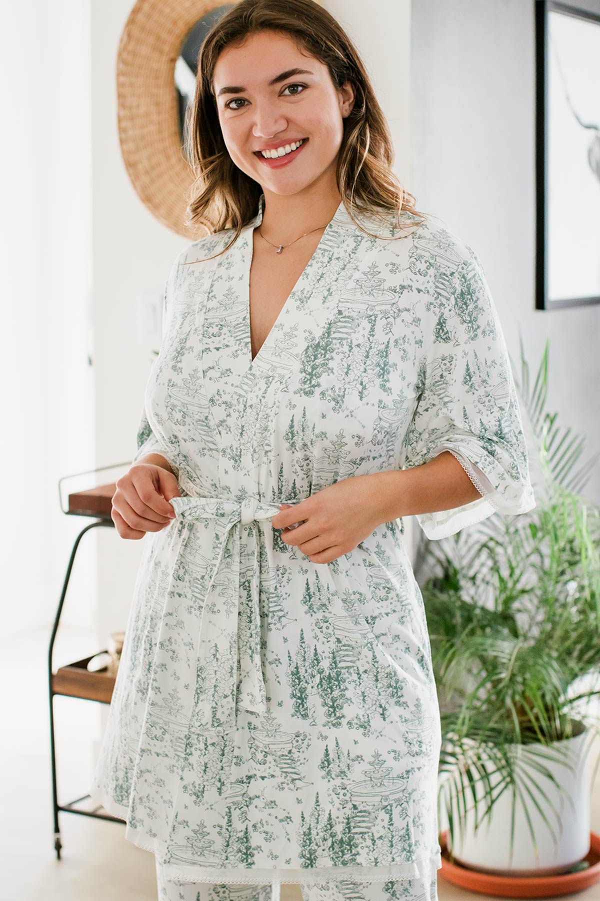 A woman standing with a smile on her face, her hands adjusting her robe's belt, wearing Iris Kimono Sleeve Belted Bamboo Lace Robe in English Garden Print