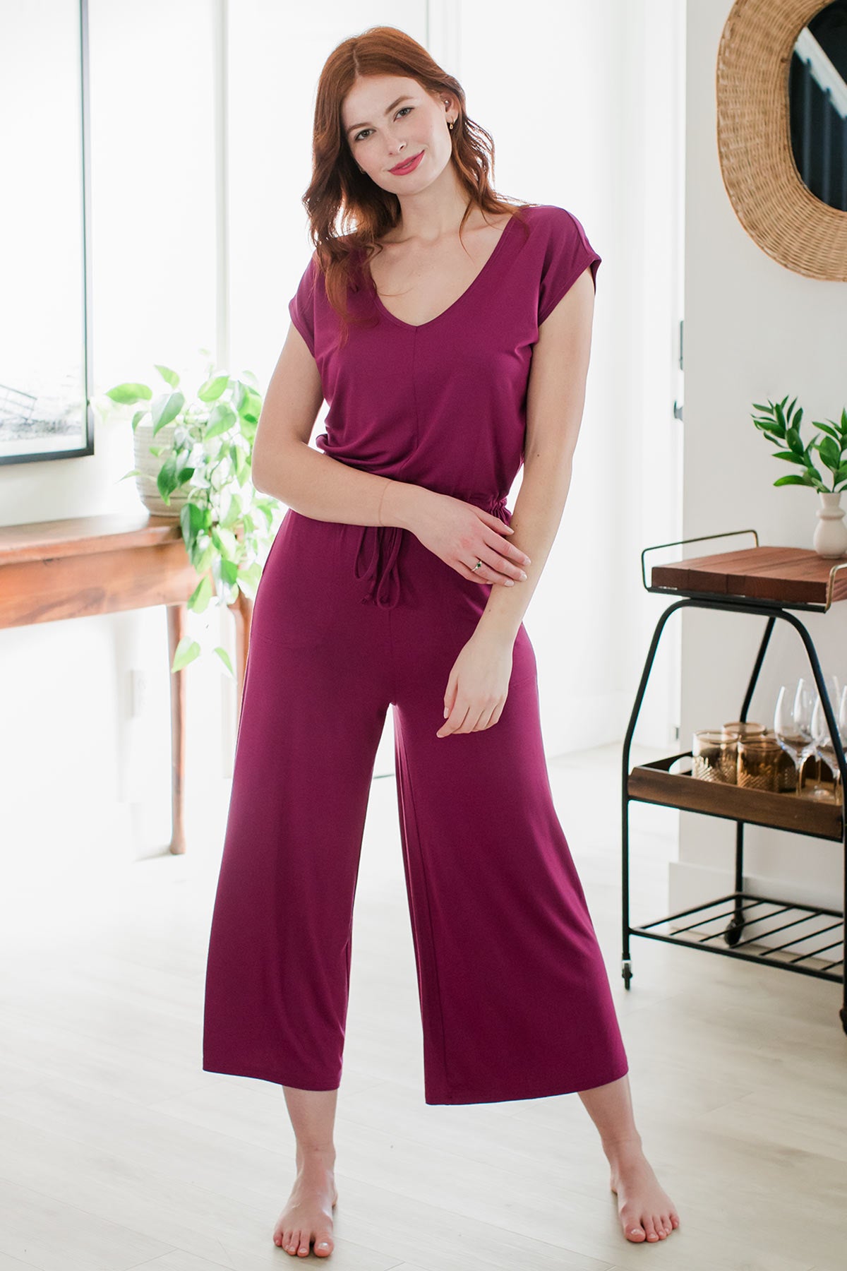 A woman standing with a wide stance and her hips tilted to the side, one hand crossed in front of her to touch her other arm, wearing Yala Kiova V-Neck Bamboo Jumpsuit in Boysenberry