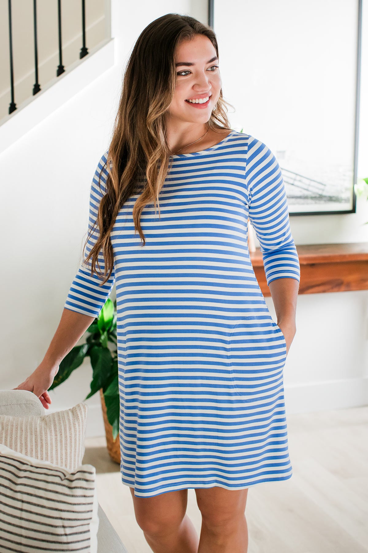 A woman standing with one hand resting on the back of a couch and her other hand in her pocket, wearing Yala Rita Boatneck A-Line Bamboo Dress with Pockets in French Blue Natural Stripe
