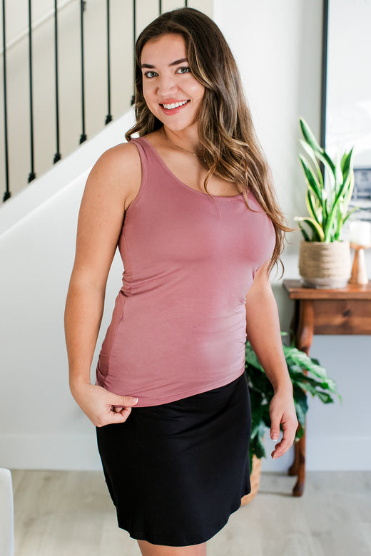 A woman standing facing to the side with one hand holding the hem of her shirt, her head turned to smile at the camera, wearing Yala Zia Layering Bamboo Tank Top in Rose