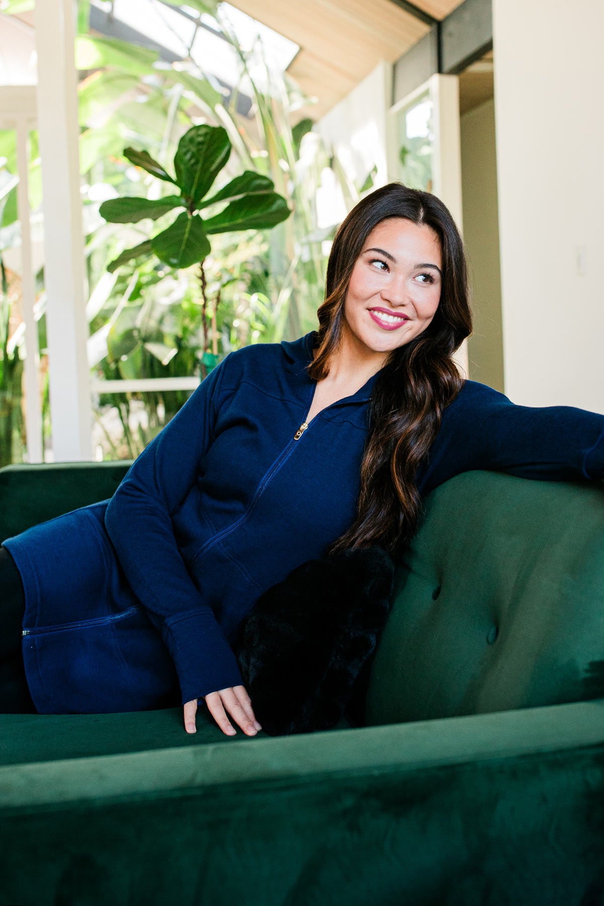 A woman sitting on a couch, on arm stretched across the back of the couch, wearing Yala Jemma Zip-Up Long Bamboo & Organic Cotton Sweatshirt Hooded Jacket in Navy