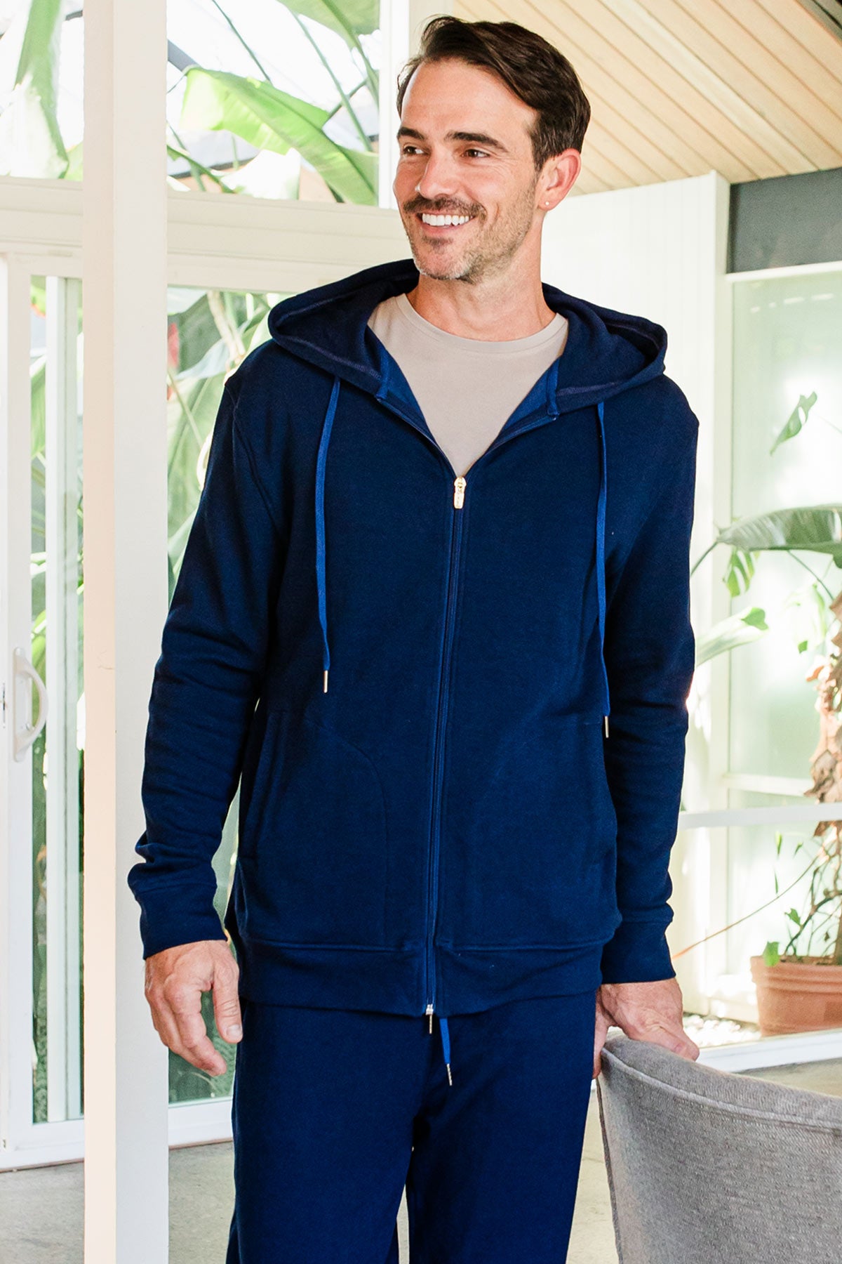 A man standing and smiling off to the side with one hand hodling on to the back of a chair, wearing Yala Men's Joey Zip-Up Bamboo & Organic Cotton Sweatshirt Hooded Jacket in Navy