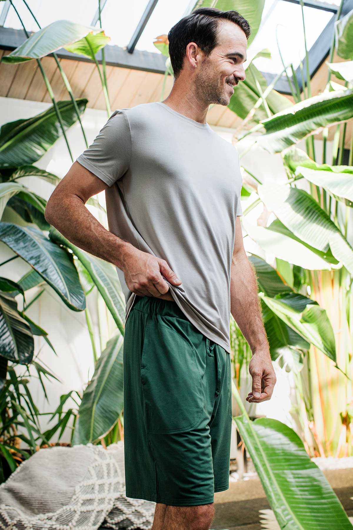 A man standing facing to the side while adjusting the hem of his shirt, a smile on his face, wearing Yala Men's Mateo ButterSoft Bamboo Lounge Shorts in Evergreen
