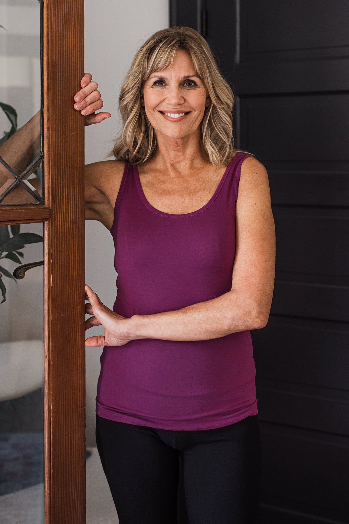 A woman standing and holding on to the frame of a glass door, wearing Yala Zia Layering Bamboo Tank Top in Boysenberry