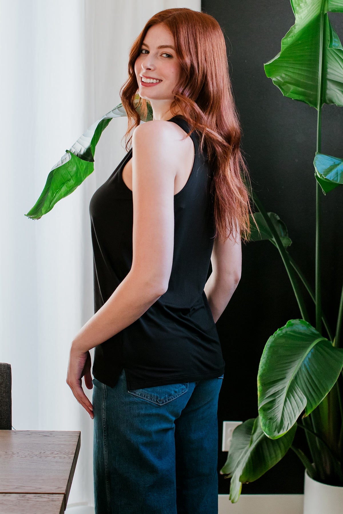 A woman standing facing away from the camera, her body turned slightly to look back towards the camera, wearing Ginger V-Neck Racerback Bamboo Tank Top in Black