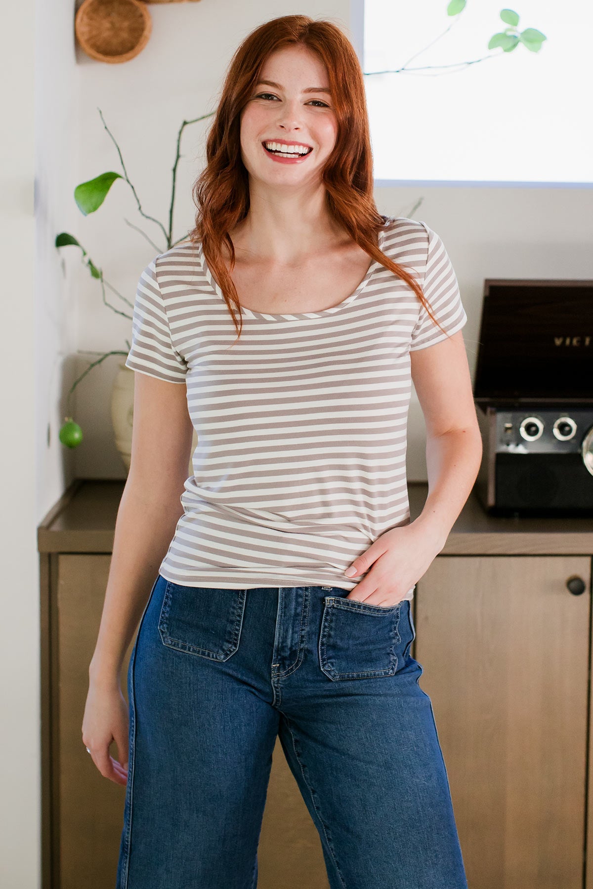 A woman standing with one hand in her pants pocket and a smile on her face, wearing Yala Heidi Scoop Neck Bamboo Tee Shirt in Ash Natural Stripe