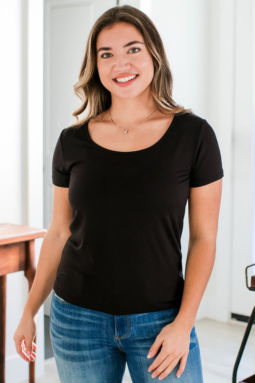 A woman standing with one hand lightly resting on the front of her thigh, wearing Yala Heidi Scoop Neck Bamboo Tee Shirt in Black