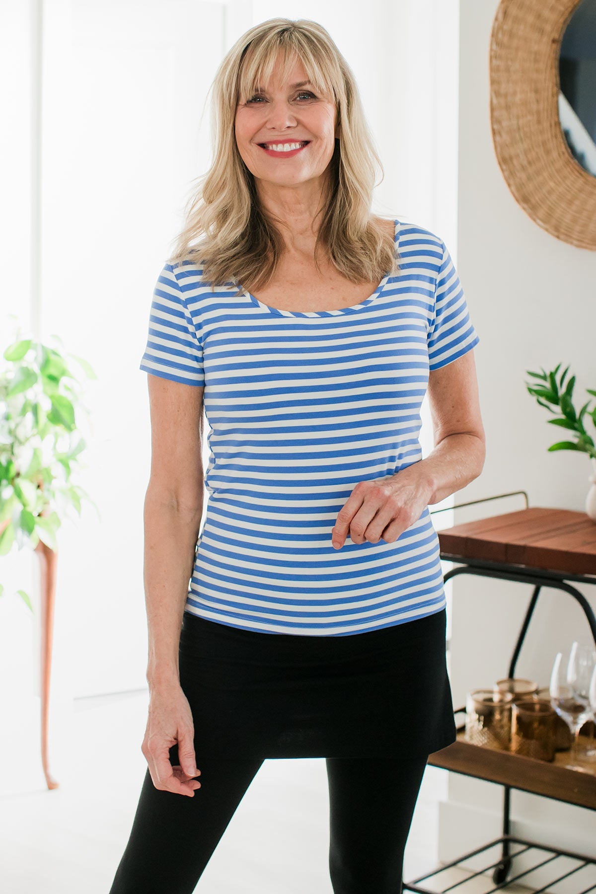 A woman standing and smiling with one hand held in front of her, wearing Yala Heidi Scoop Neck Bamboo Tee Shirt in French Blue Natural Stripe