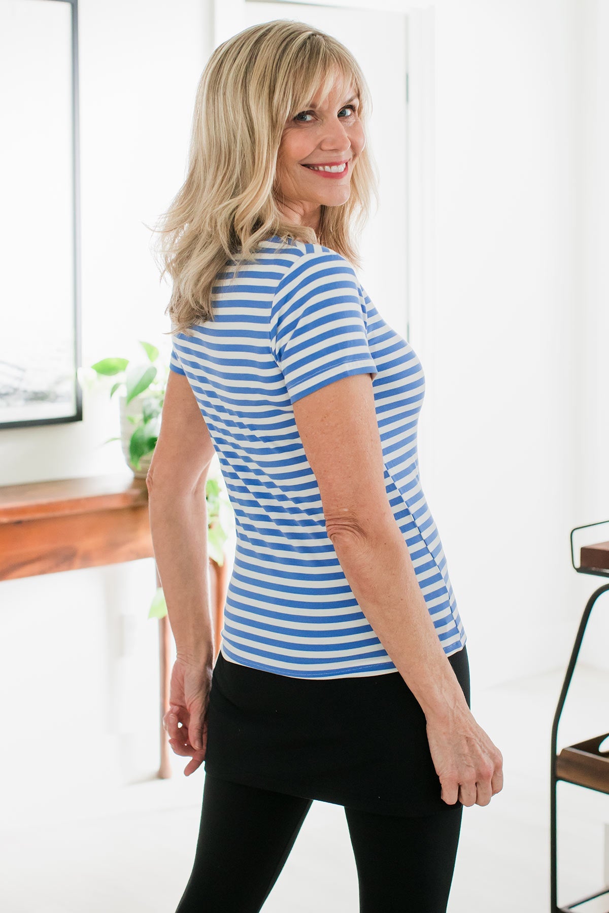 A woman standing facing away from the camera with her hands at her sides, looking back over her shoulder, wearing Yala Heidi Scoop Neck Bamboo Tee Shirt in French Blue Natural Stripe