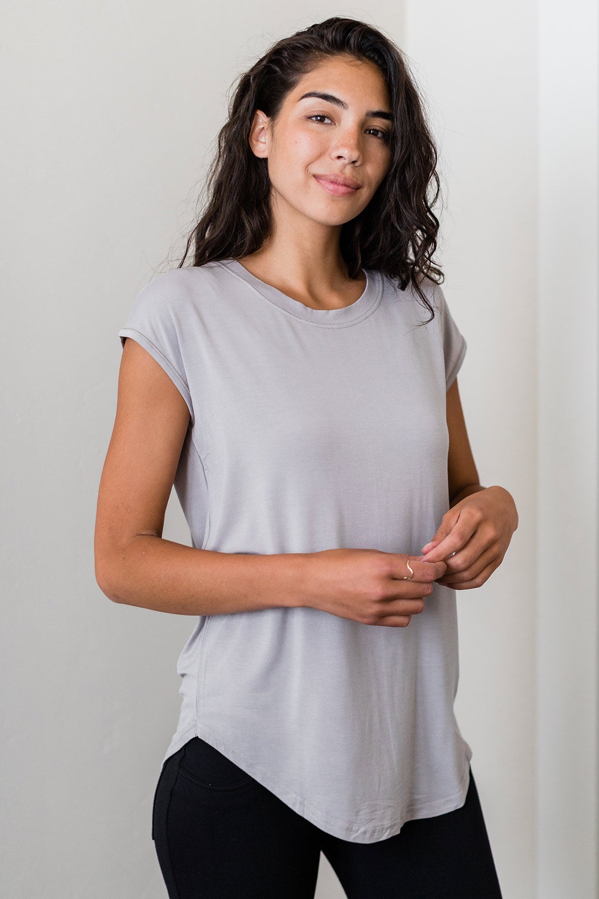 A woman standing and smiling with both hands held in front of her, wearing Yala Cassidy Cap Sleeve Bamboo Boyfriend Tunic in Ash