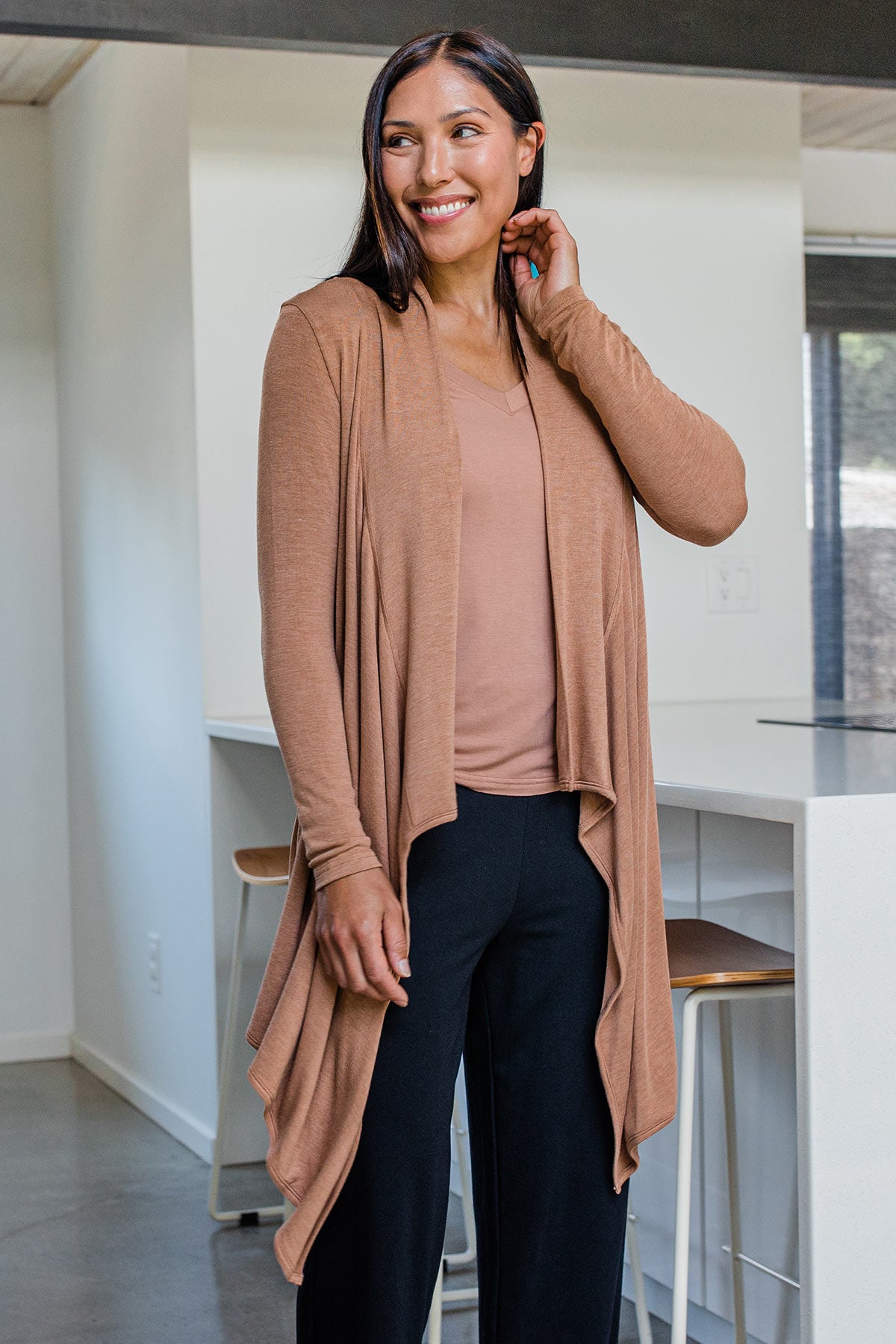 A woman standing and smiling while looking to the side, wearing Yala Hilary Bamboo Sweater Wrap in Camel