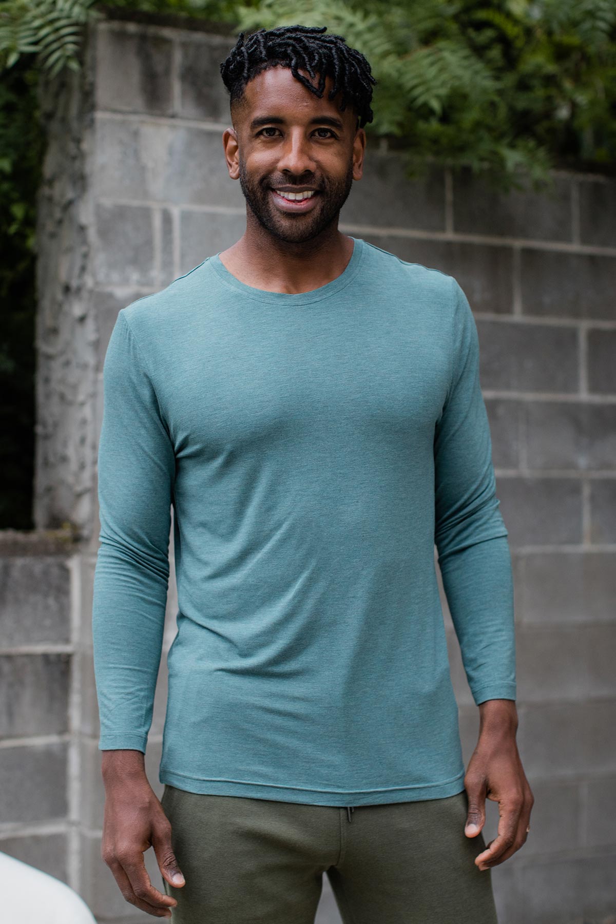 A man standing and smiling with his hands at his sides, wearing Yala Jonah Men's Long Sleeve Bamboo Crew Tee Shirt in Teal Melange