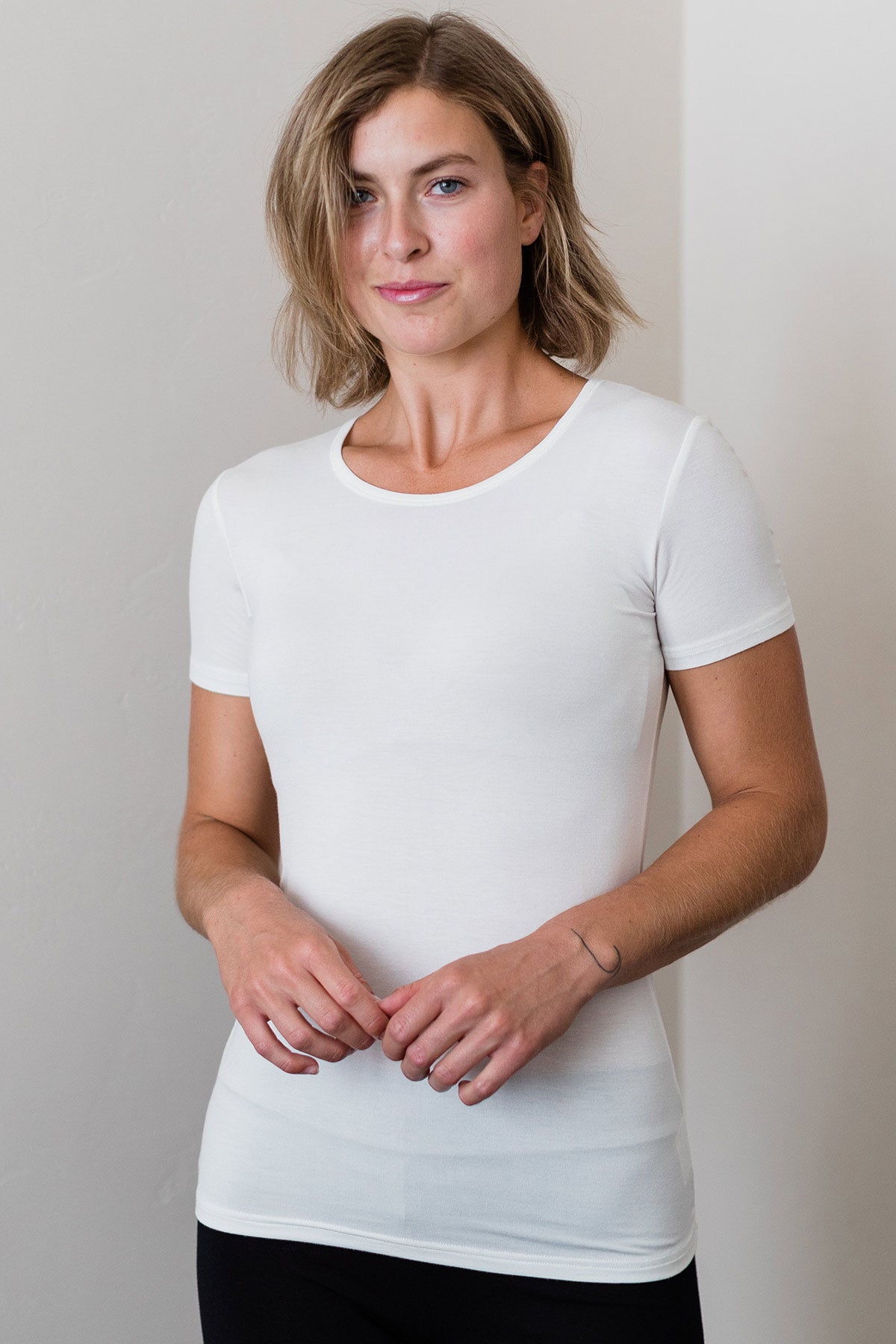 A woman standing with both hands held in front of her waist, wearing Yala Nicole Short Sleeve Fitted Bamboo Tee Shirt in Natural