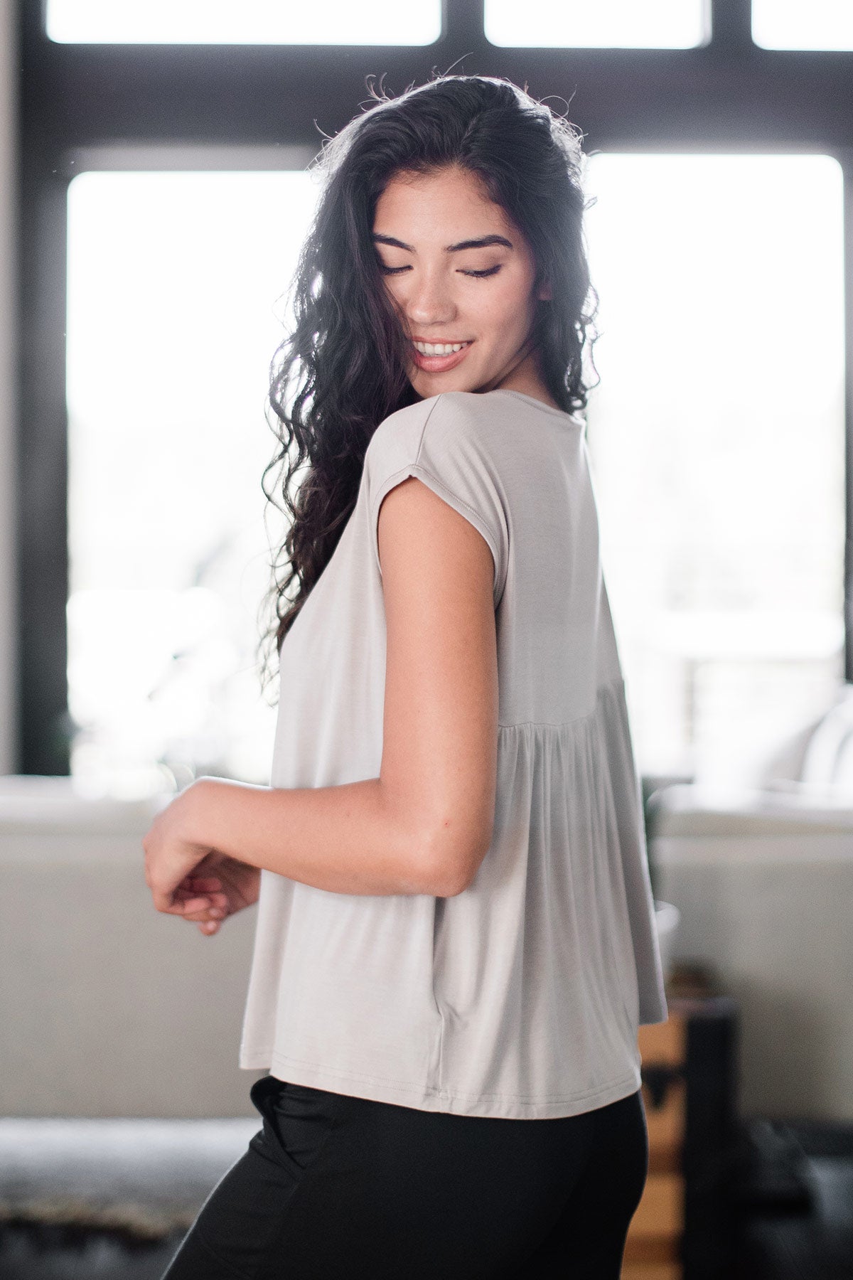 A woman standing facing away from the camera and turning to look over her shoulder, wearing Yala Opal Swing Bamboo Top in Ash