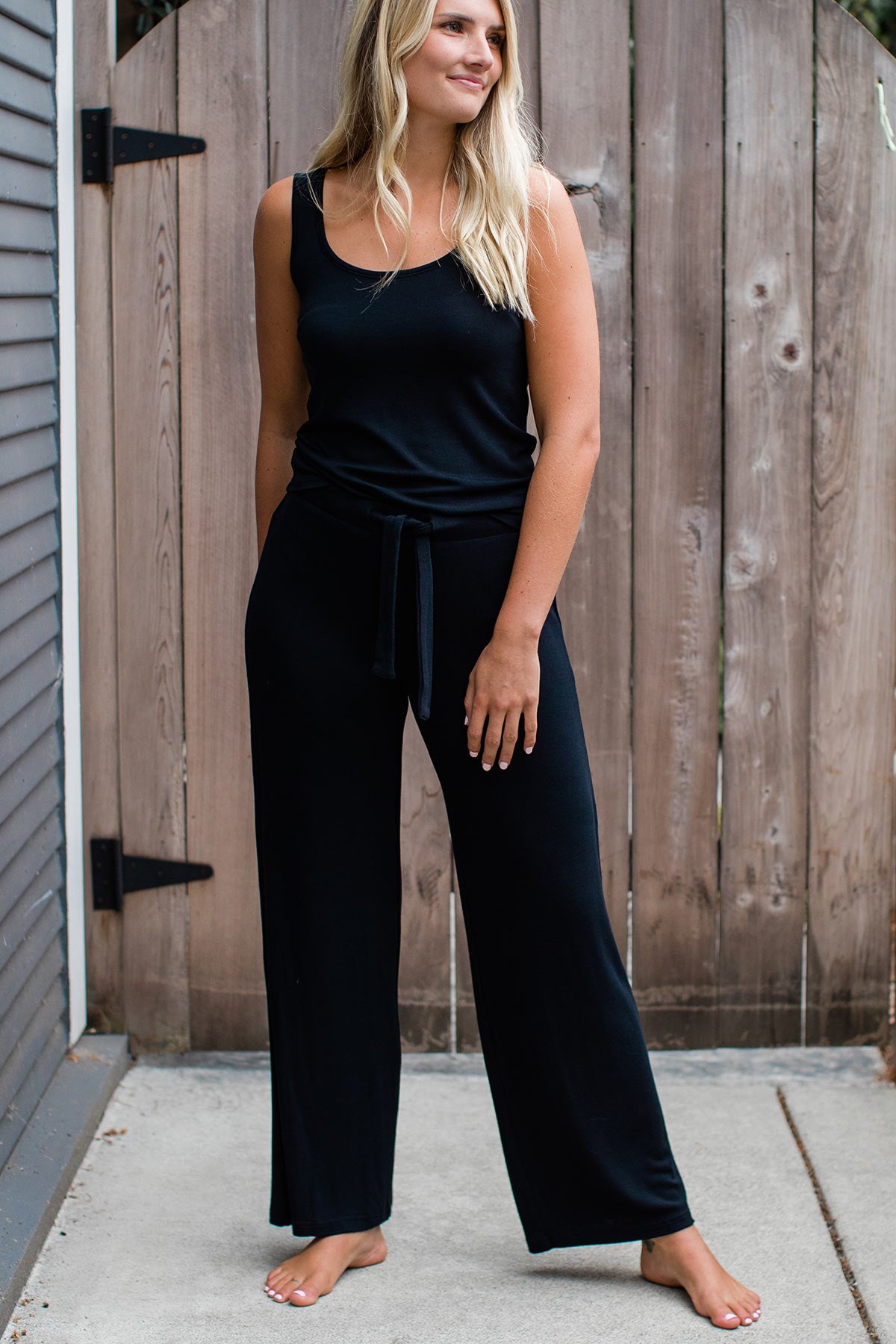 A woman standing and smiling with one leg stretched out to the side, wearing Yala Peyton Bamboo Sweater Drawstring Lounge Pants in Black