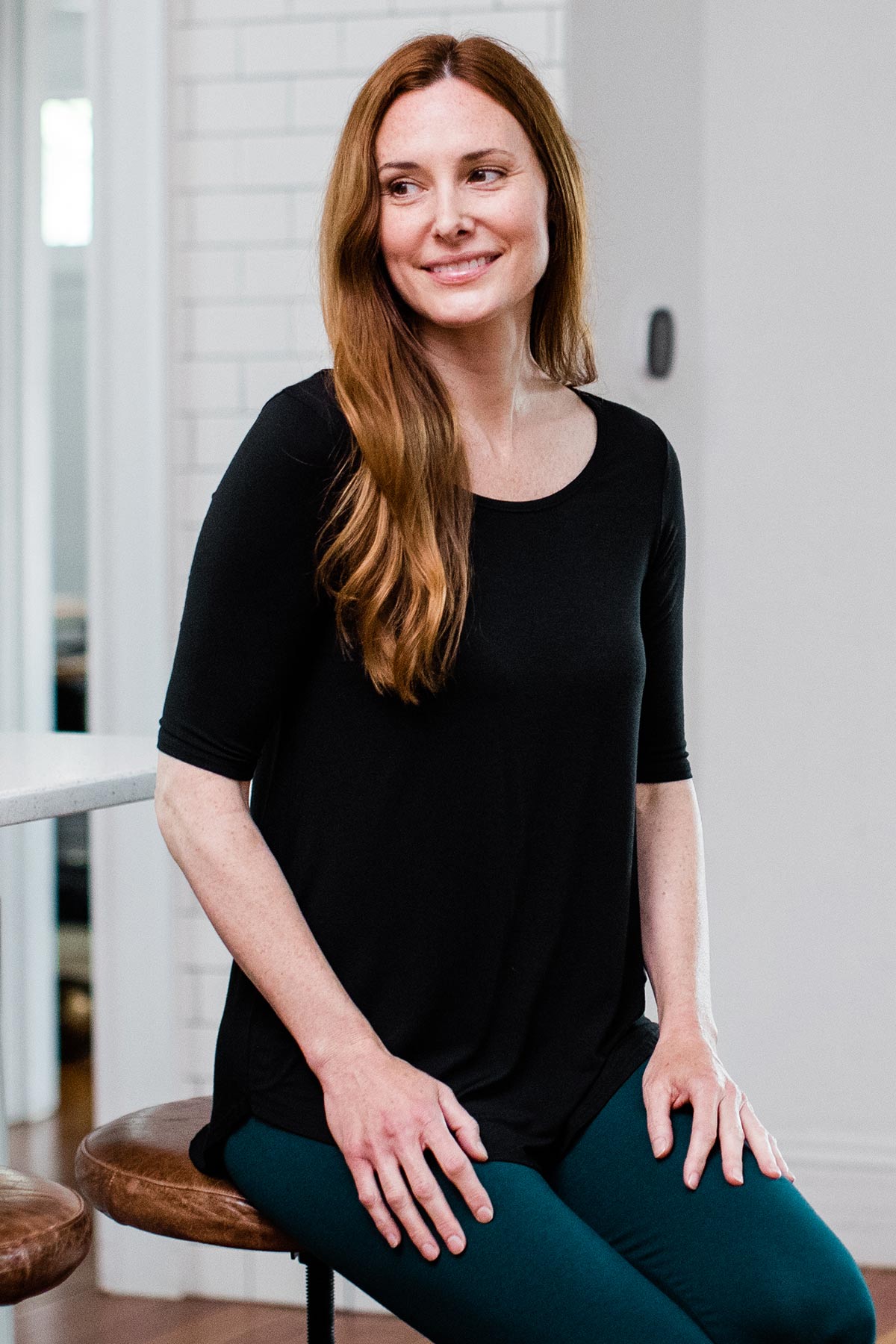 A woman sitting on a stool and looking to the side while smiling, wearing Yala Sandy Relaxed Fit Scoop Neck Short Sleeve Bamboo Top in Black