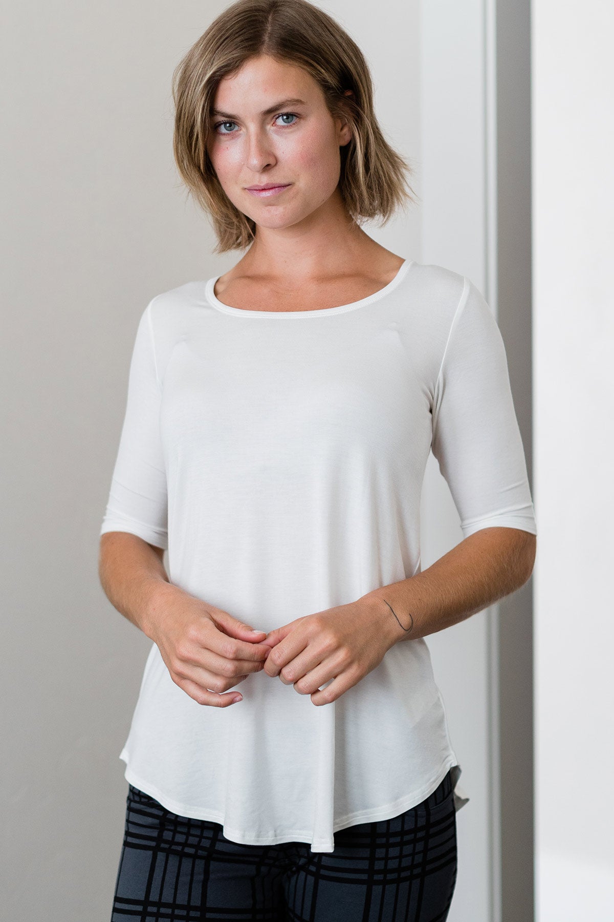 A woman standing with both hands held in front of her waist, wearing Yala Sandy Relaxed Fit Scoop Neck Short Sleeve Bamboo Top in Natural
