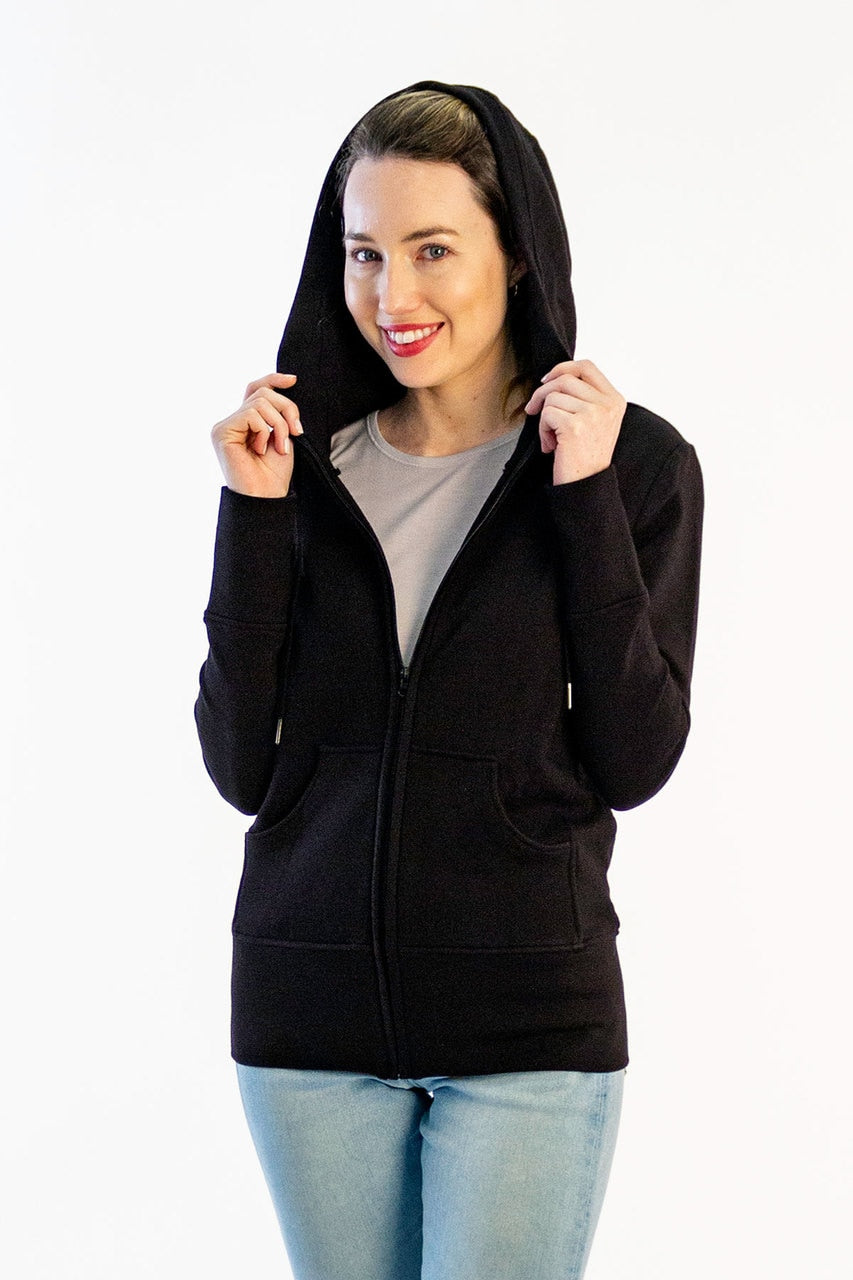 A woman standing and smiling with her sweater hood up and booth hands in the hood's edge, wearing Yala Scarlet Zip-Up Bamboo and Organic Cotton Hooded Jacket Sweatshirt in Black
