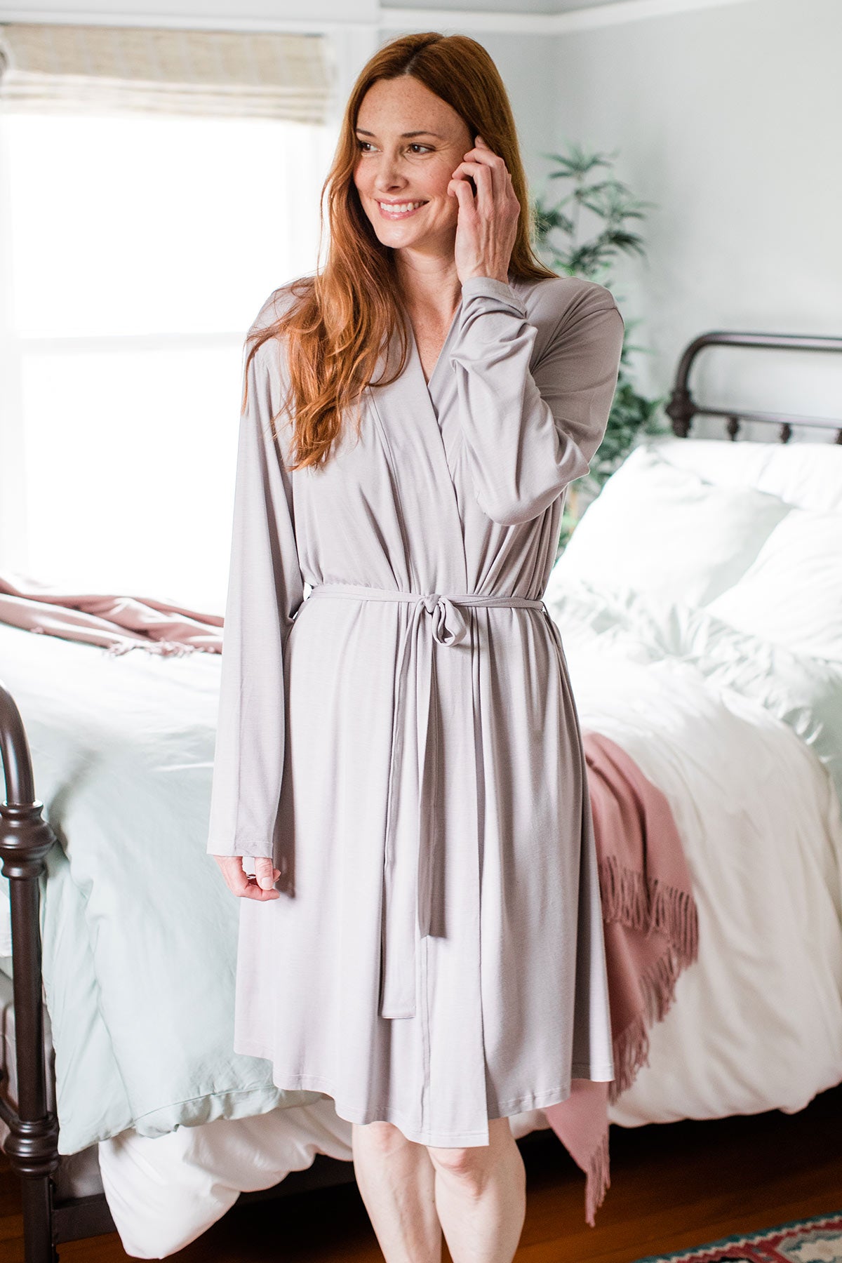 A woman standing and smiling while brushing her hair aside with one hand, wearing Yala Serenity Long Sleeve Short Belted Bamboo Robe in Ash