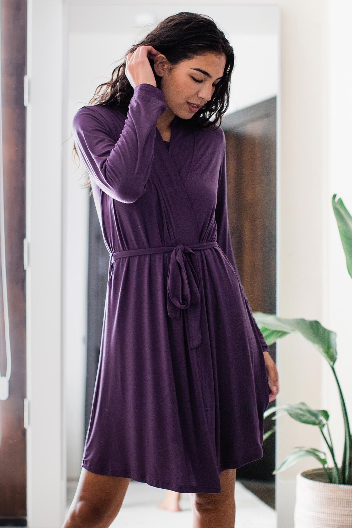 A woman standing and looking downward while running one hand through her hair, wearing Yala Serenity Long Sleeve Short Belted Bamboo Robe in Aster