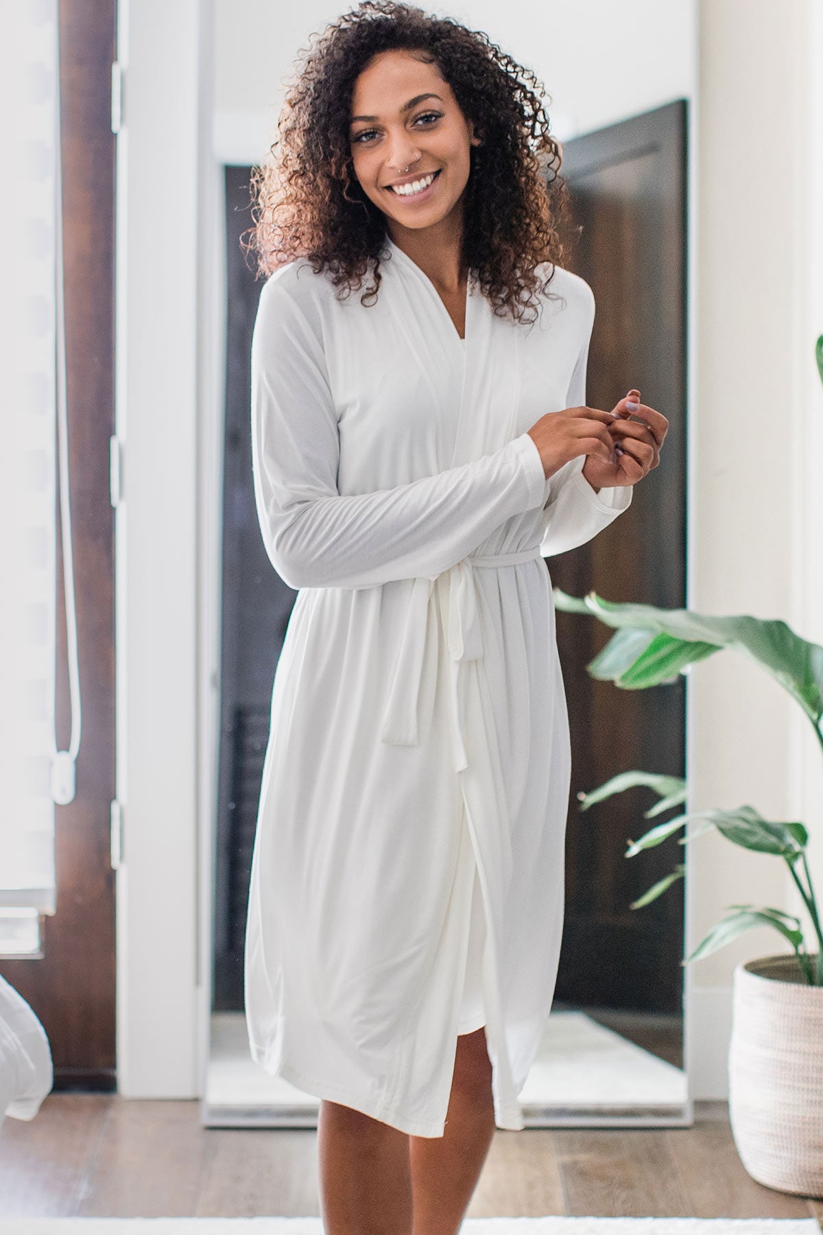 A woman standing and smiling with both hands held to one side, wearing Yala Serenity Long Sleeve Short Belted Bamboo Robe in Natural