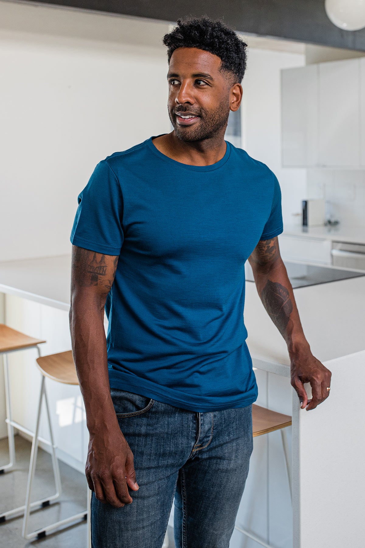 A man standing and leaning against a countertop, looking to the side and wearing Yala Superfine Merino Wool Short Sleeve Tee in Lapis