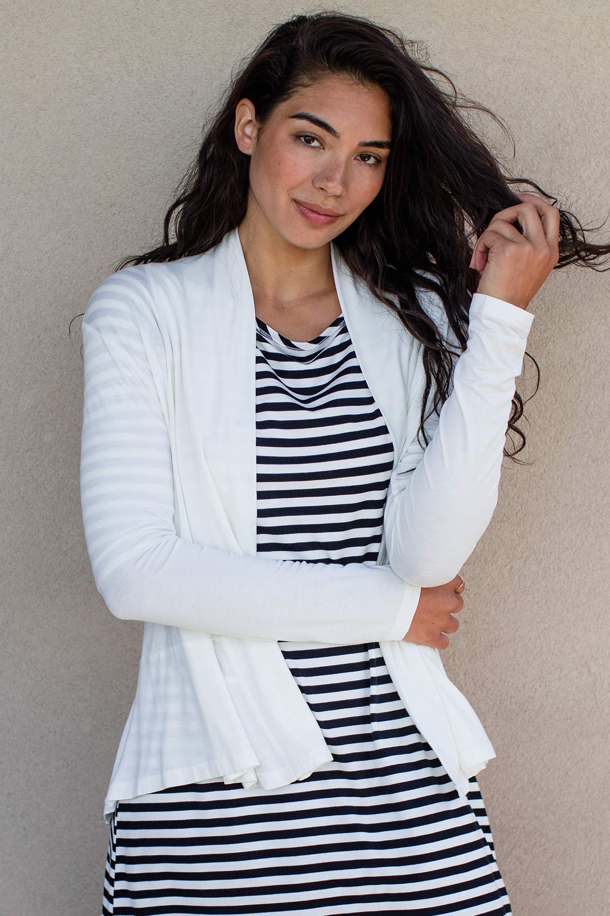 A woman standing with one hand holding her waist and the other in her hair, wearing yala Tatum Hi-Low Bamboo Cardigan Wrap in Natural