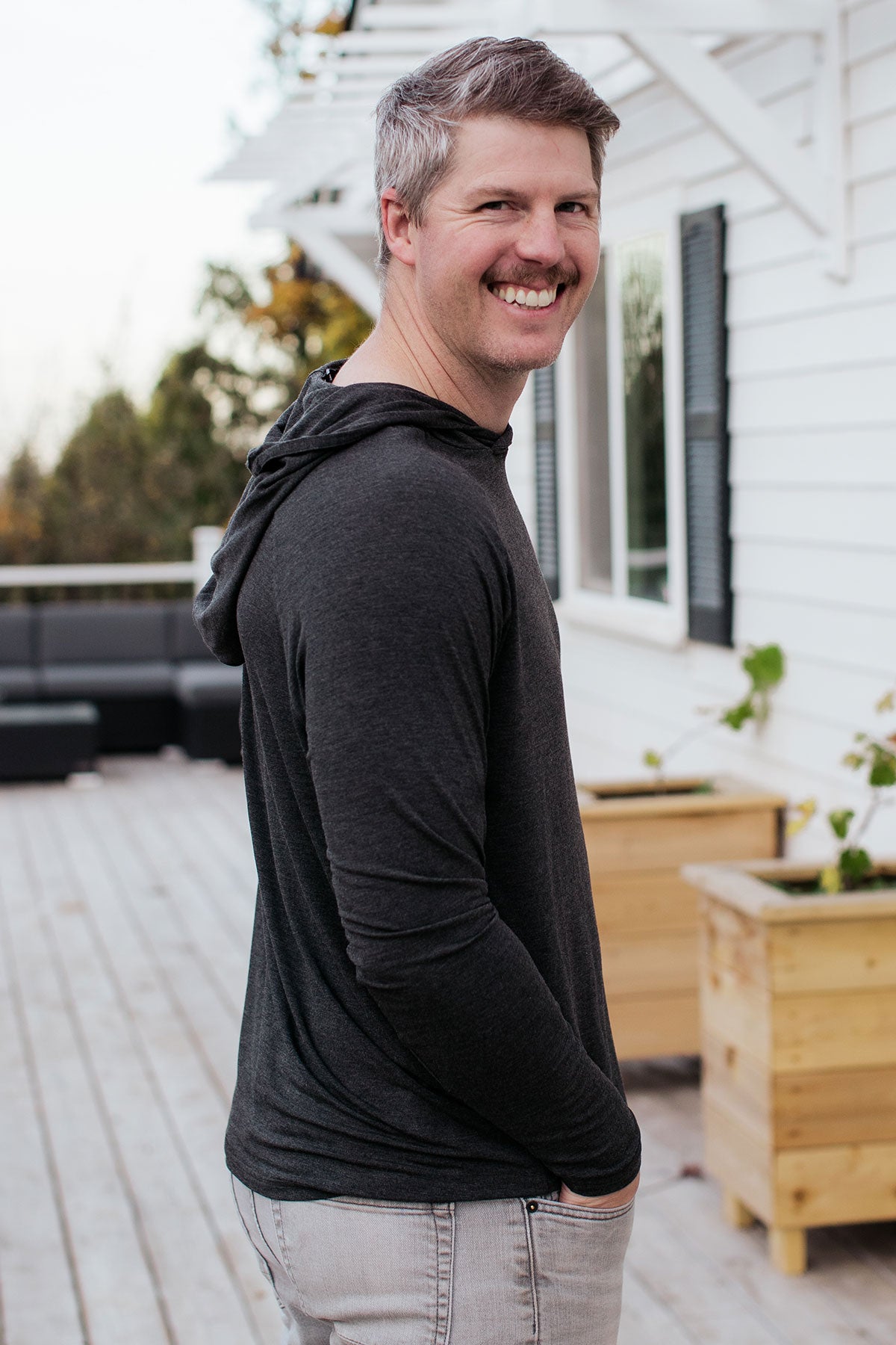 A man standing facing sideways and smiling towards the camera with both hands in his pockets, wearing Yala Travis Men's Hooded Long Seeve Raglan Bamboo Tee Shirt in Charcoal Melange