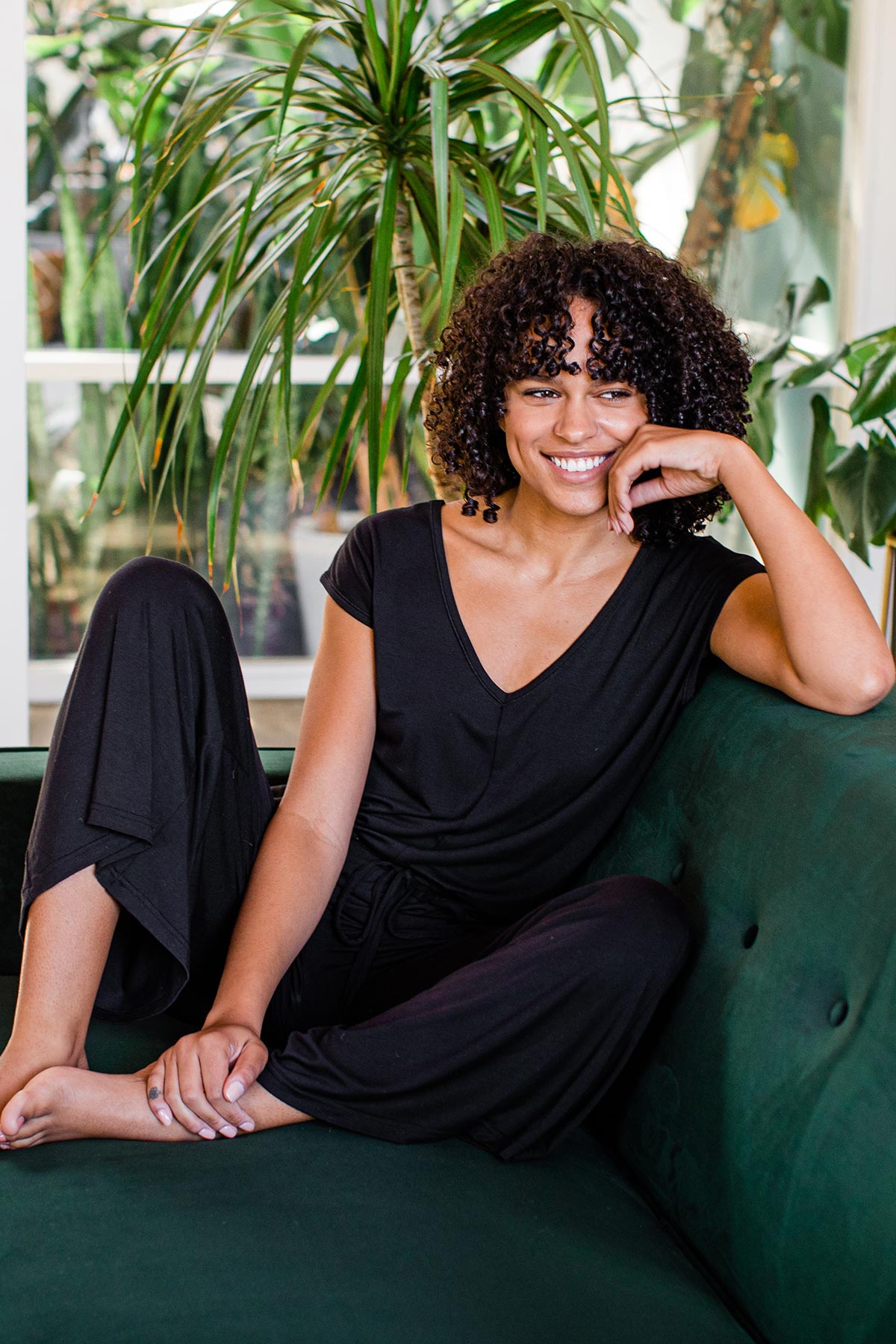 A woman sitting on a couch and smiling, wearing Yala Kiova V-Neck Bamboo Jumpsuit in Black
