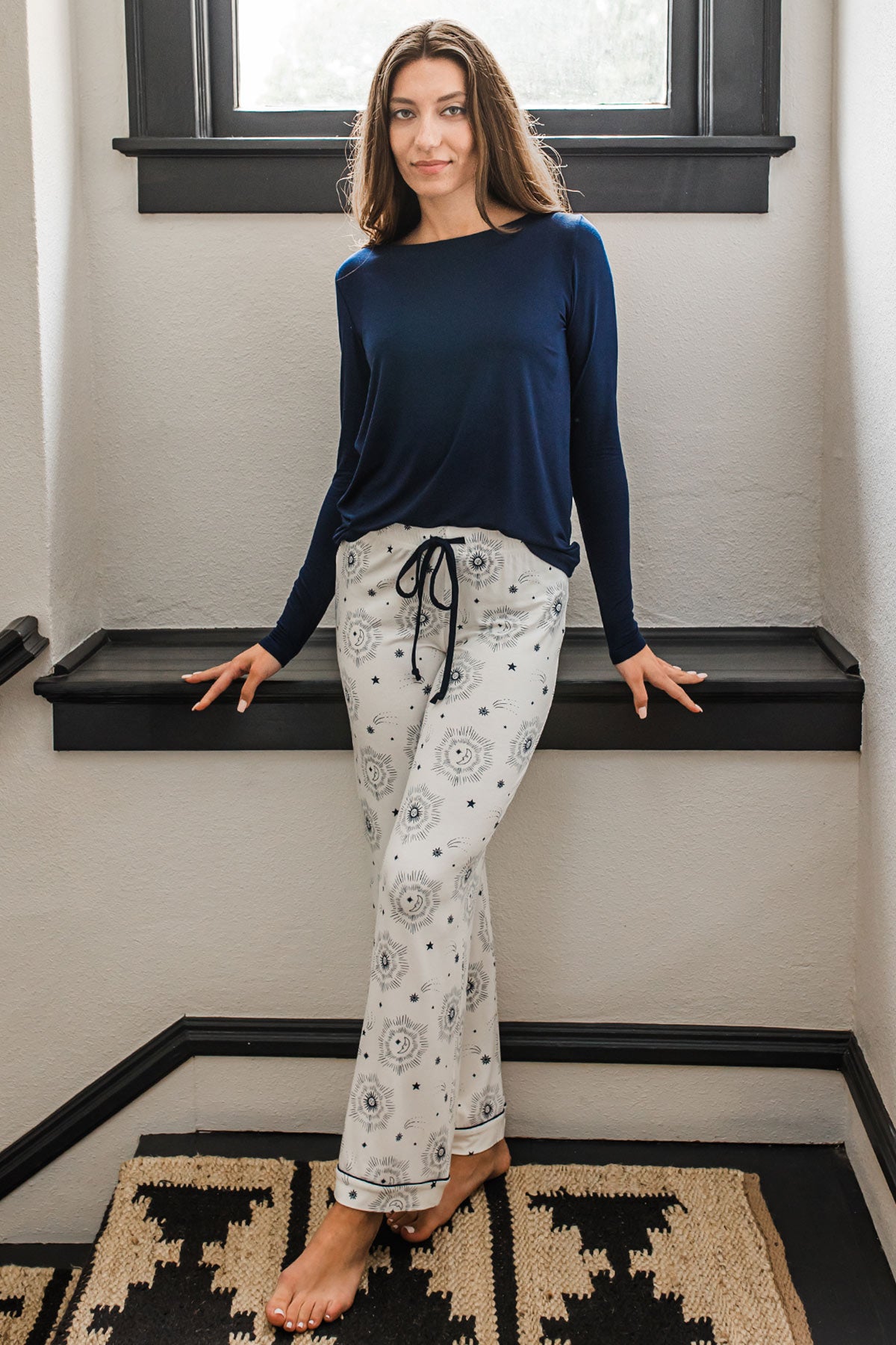 A woman standing in a stairwell and leaning back against a shelf, wearing Yala Gillian Piped Bamboo Pajama Pants